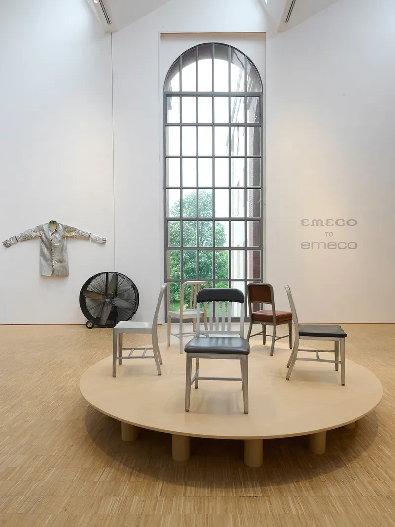 Emeco chairs on a round platform in a gallery space with an arched window