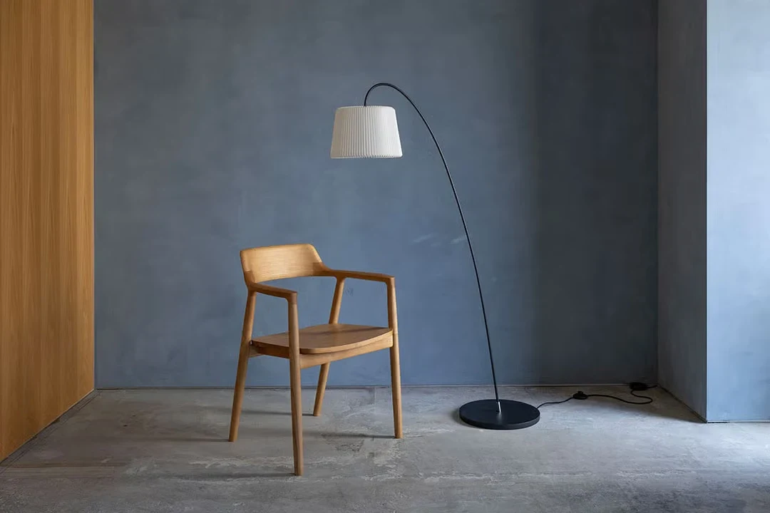 A wooden armchair with a curved arm rail beside an arched floor lamp against a plain wall