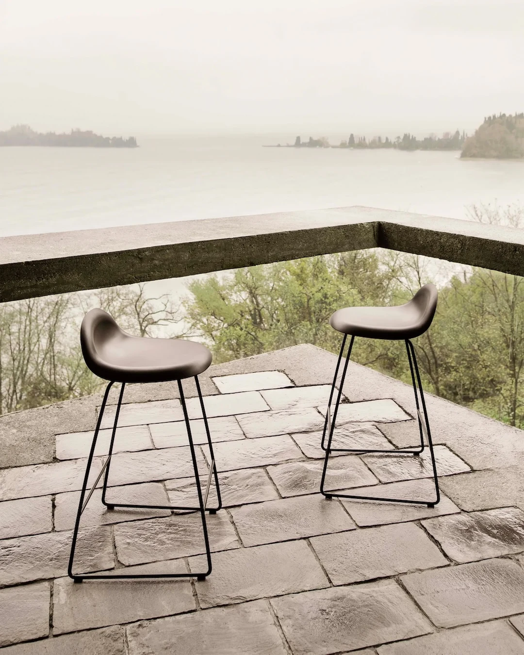 Two stools with slim legs outdoors, with water and trees behind