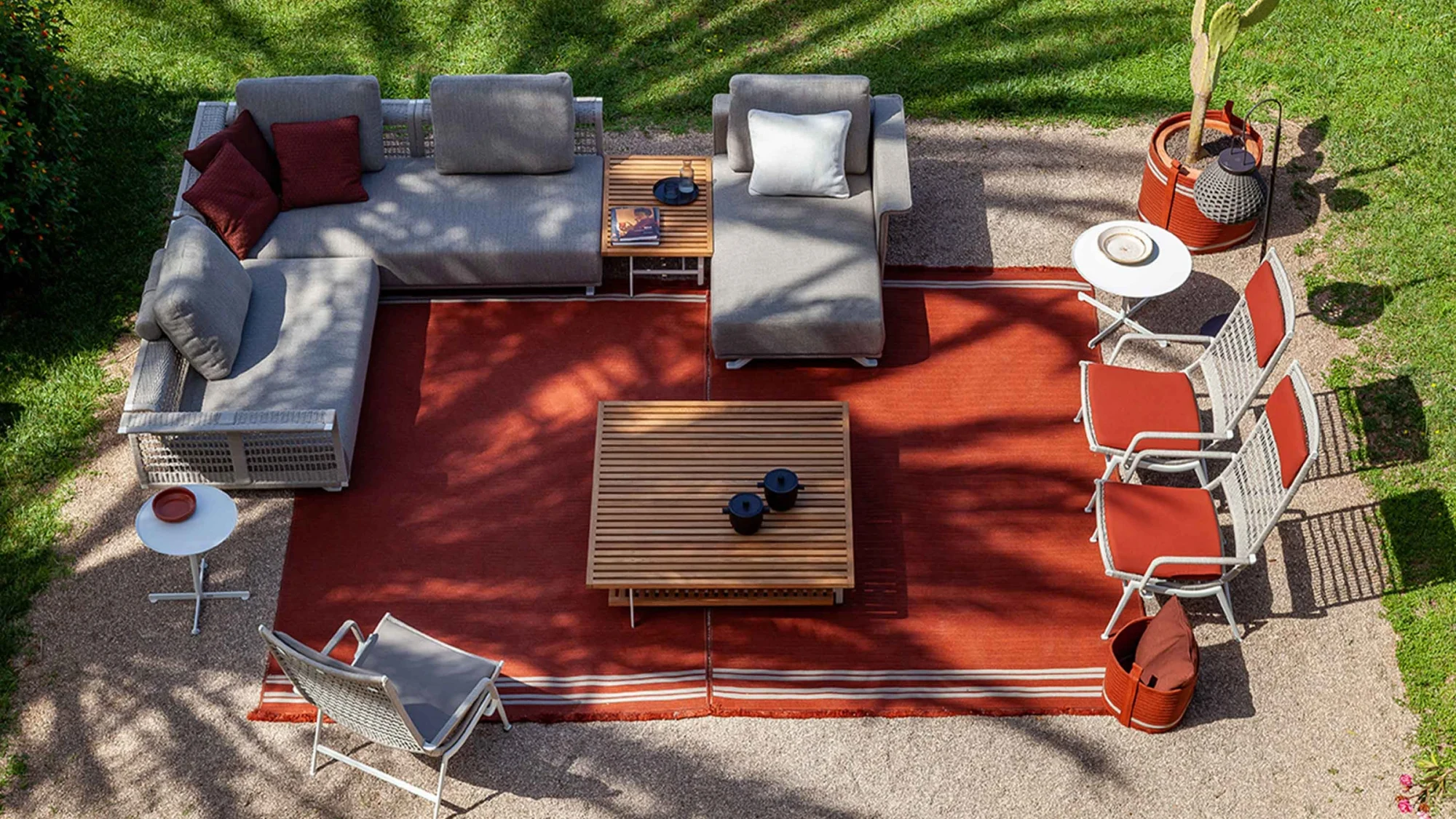 Aerial view of an outdoor corner seating composition on a red rug with a low table, side tables and armchairs