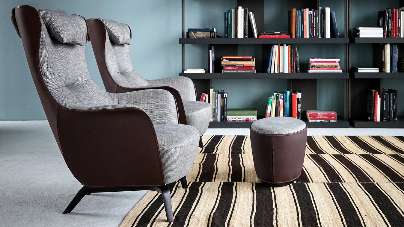 Two Mamy Blue armchairs and a round ottoman on a striped rug in front of shelves with books