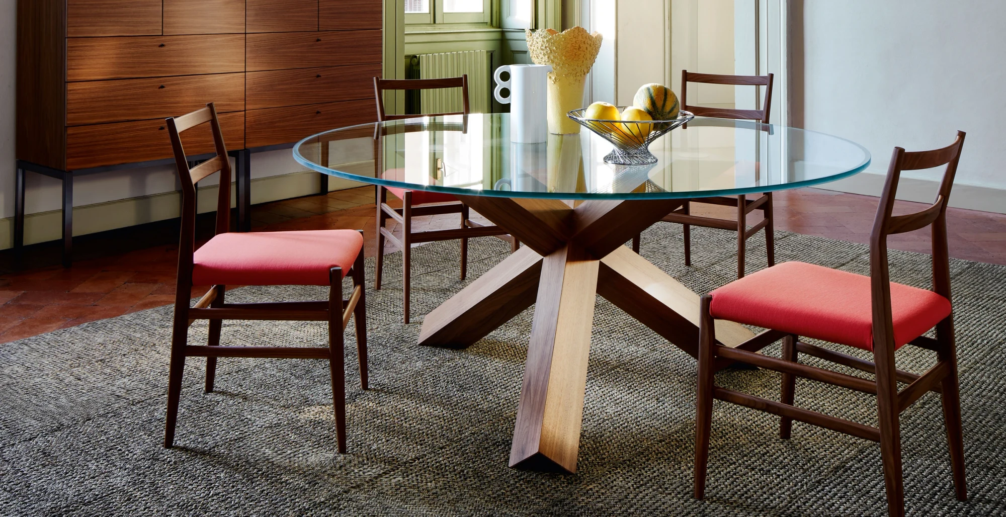 A round glass-topped table on a crossed wooden base in a room with chairs