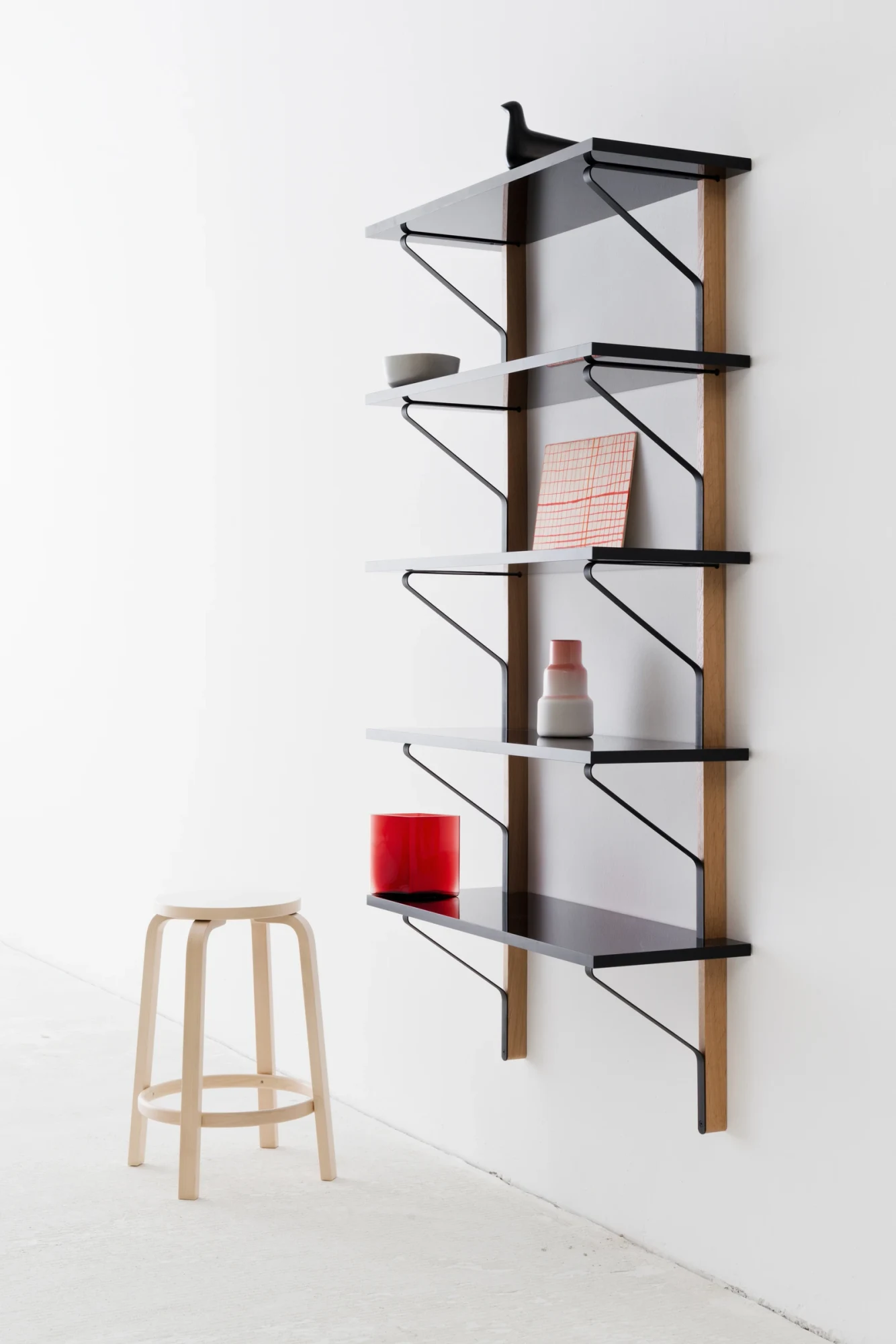 Kaari Wall Shelf 作为一个高而窄的单元安装在墙面上,旁边放着一把凳子