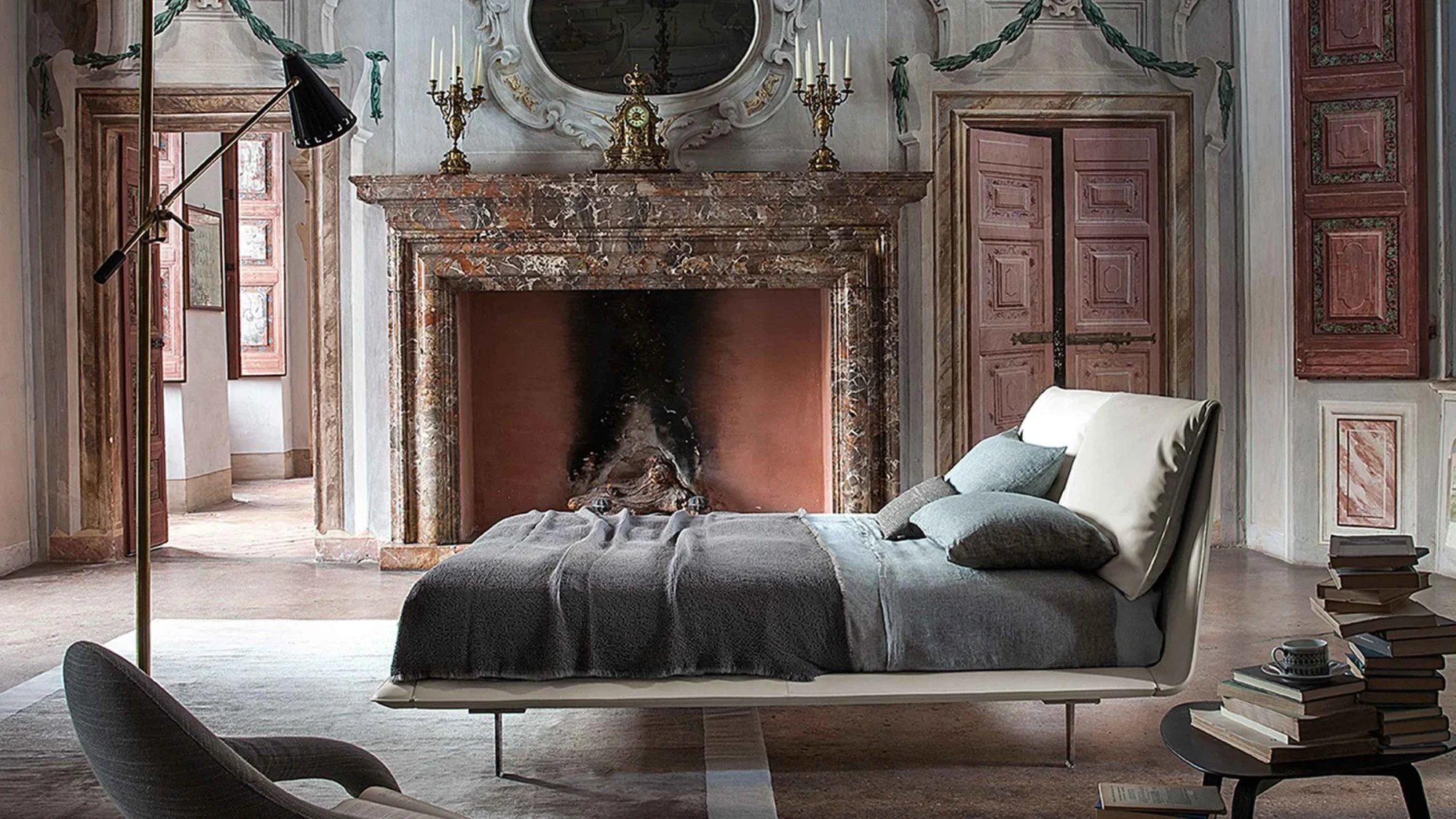 Bed with a headboard in a room with a fireplace, a floor lamp and a stack of books