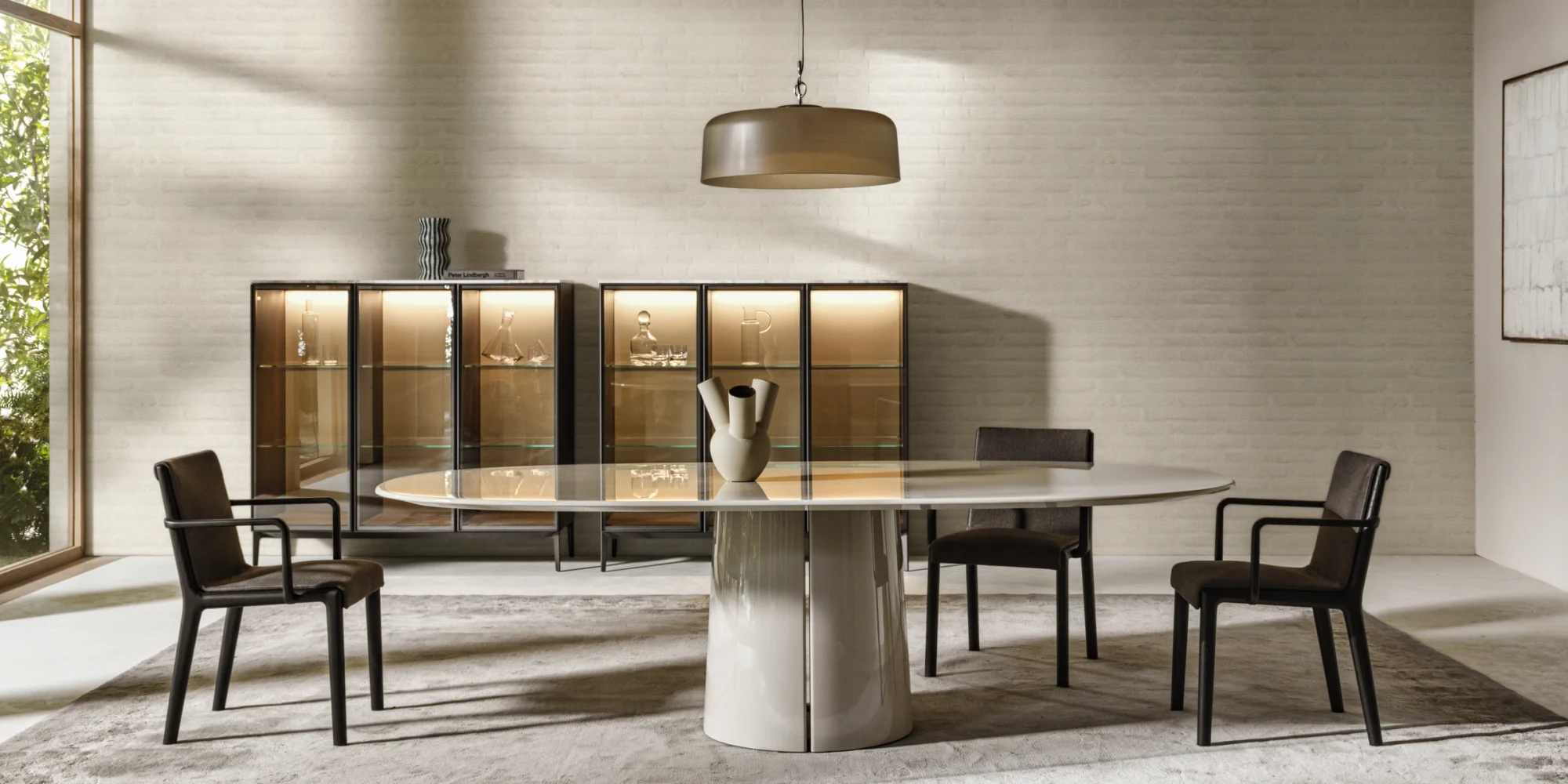 Dining table with a central base and armchairs around it, in front of a cabinet with lit shelves