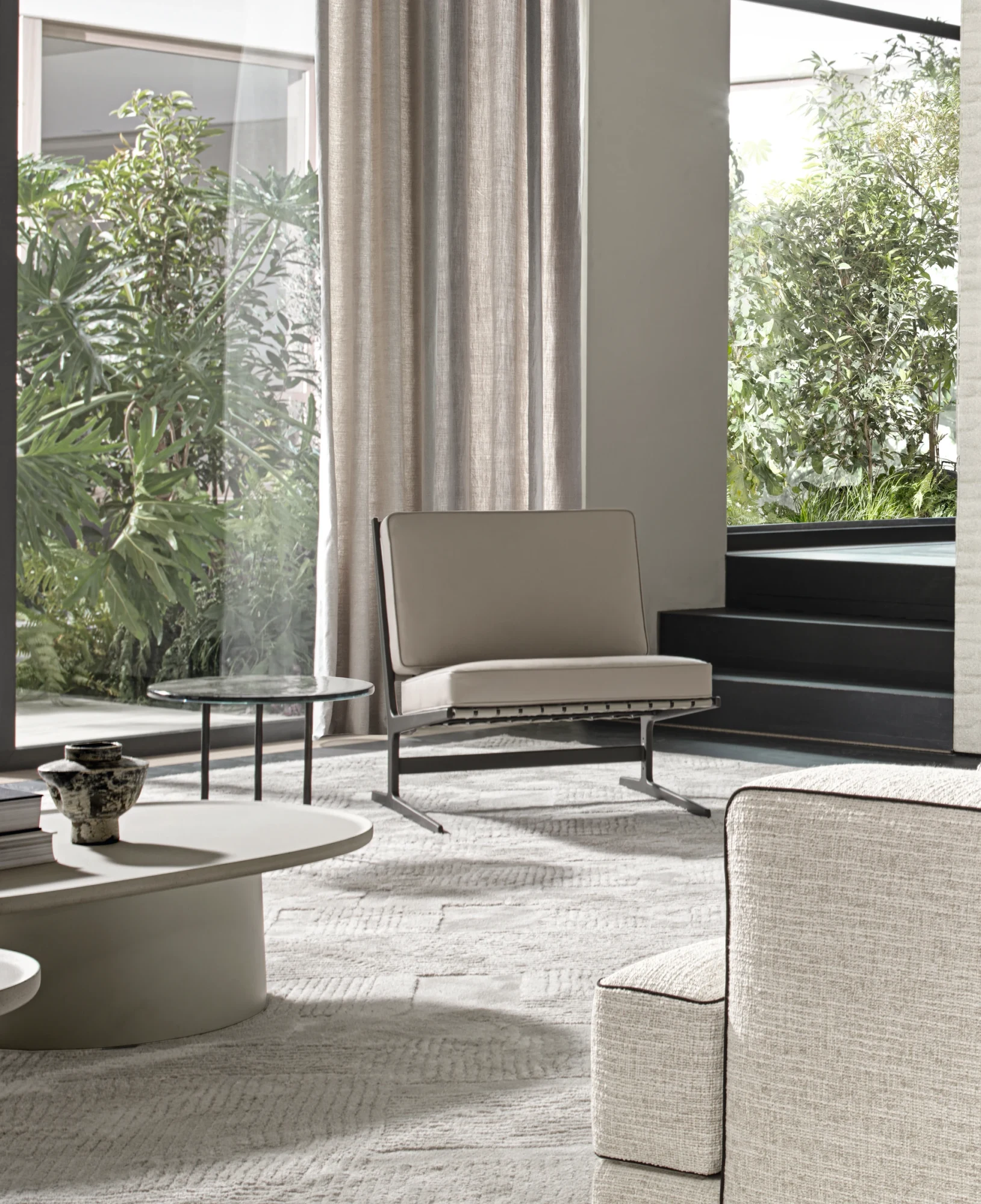 A Paula armchair in a living room with large windows, a round low table and the edge of a sofa