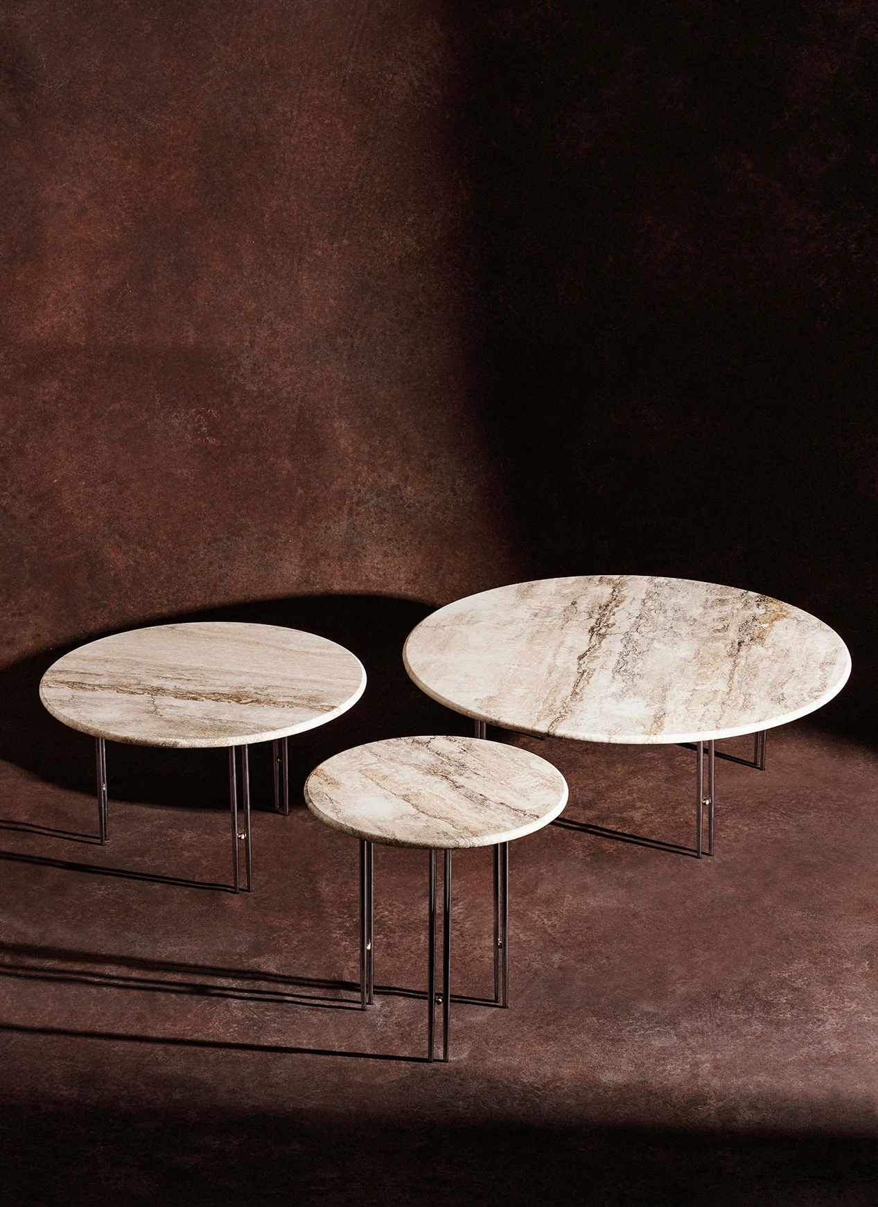 Three round tables of different sizes with pale tops and thin legs, on a dark brown background