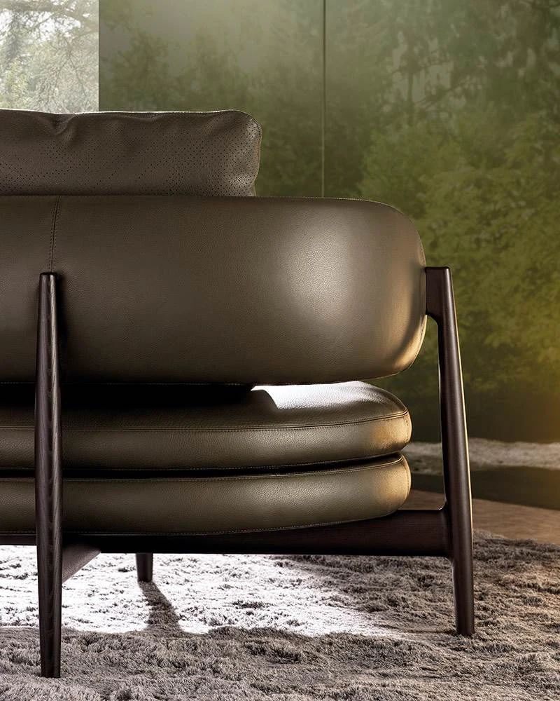 Close view of the rounded seat and back of a dark leather sofa beside a wooden leg, official Minotti photo