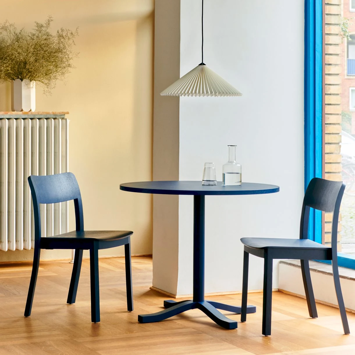 Round table with two chairs, a glass and a bottle on the tabletop and a pleated pendant lamp above it, beside a window
