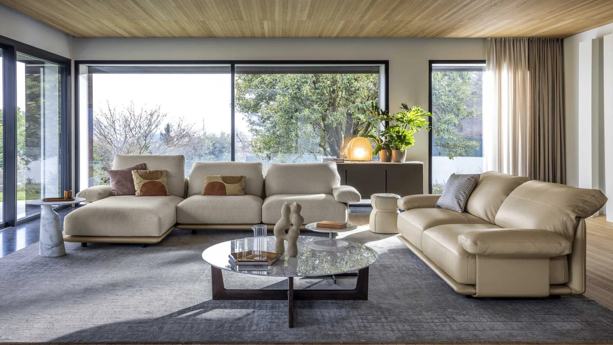 Wohnzimmer mit einem modularen Sofa, einem Chaiselongue-Ende und einem runden niedrigen Tisch vor großen Fenstern