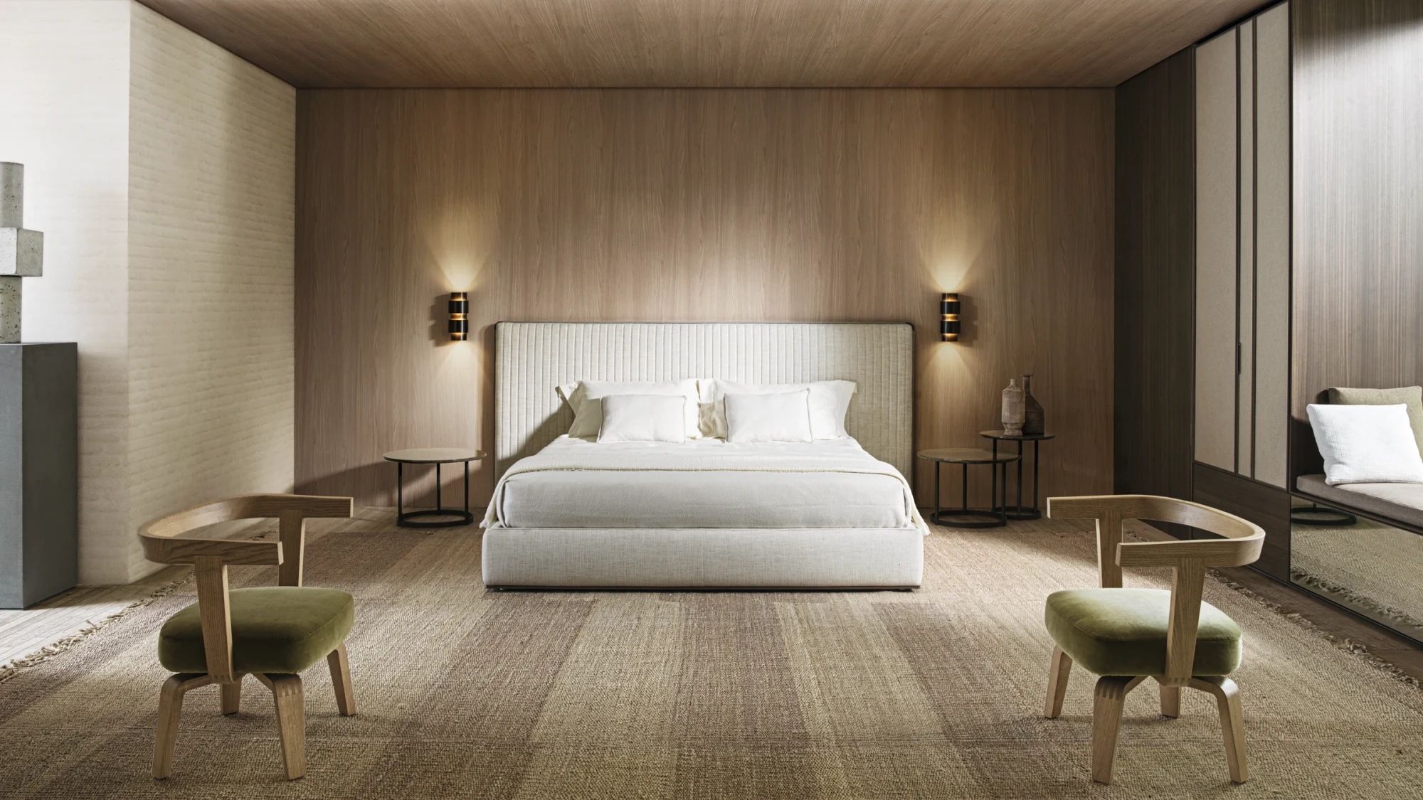 A bed with a tall rectangular headboard between two wall lamps, with two armchairs in the foreground