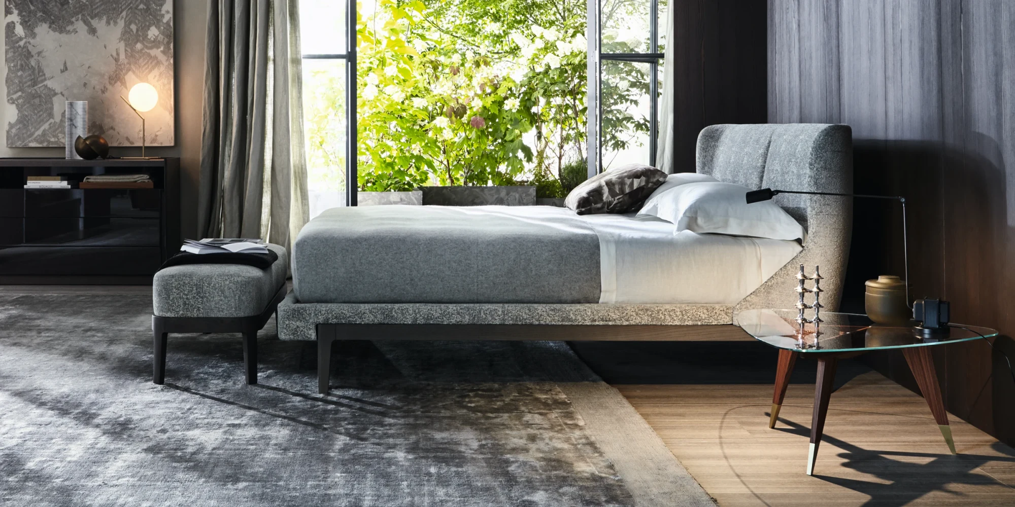 A bed with a wrap-around headboard and a low seat at its foot, in a room with a window and trees