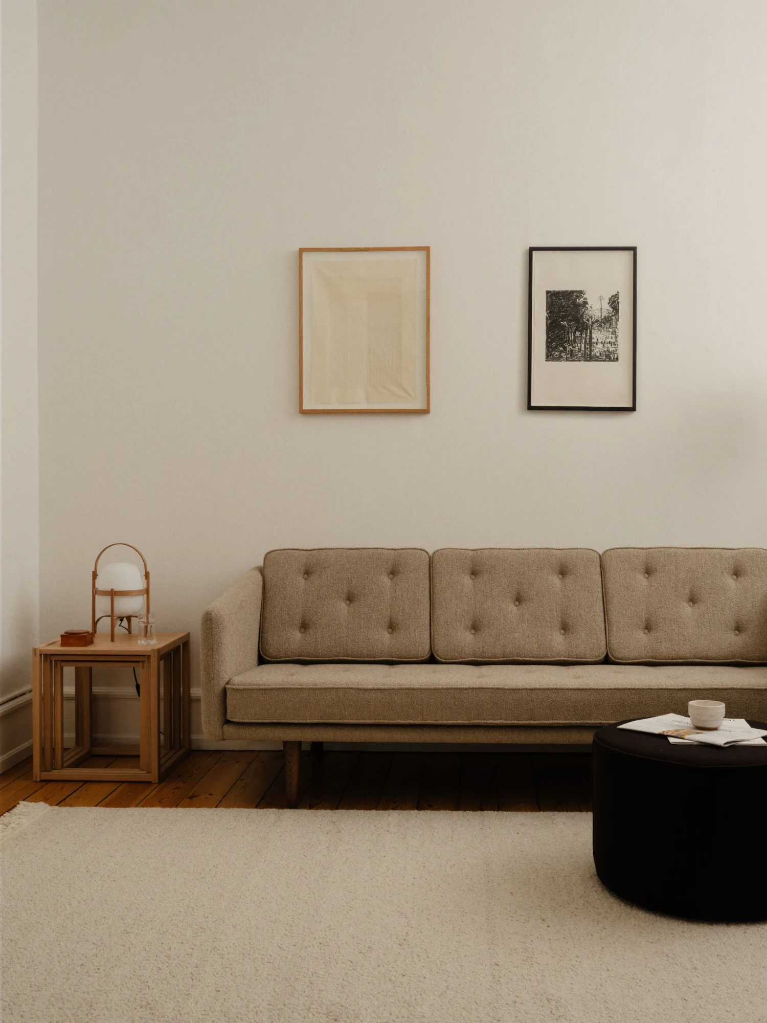 A sofa along a wall with two framed pictures, a small table at the left and a round pouf in front