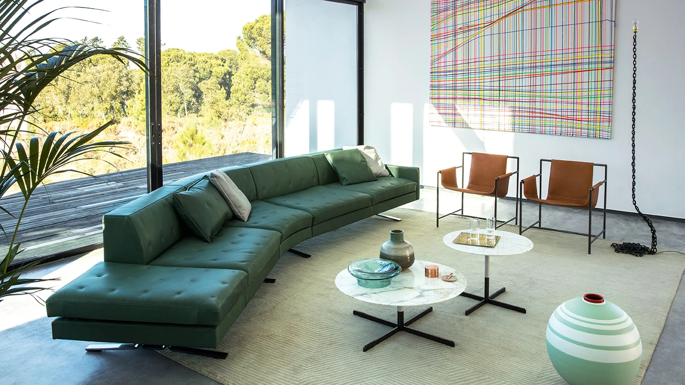 Green sofa with a chaise end and round low tables in a room with floor-to-ceiling windows and chairs